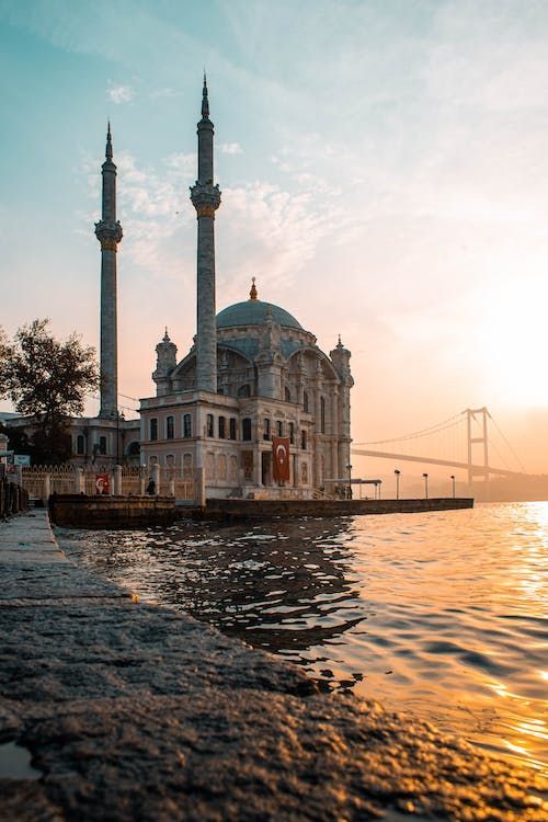Una mezquita ornamentada con dos minaretes se alza junto al agua al atardecer, con un puente a lo lejos y reflejos en el agua.