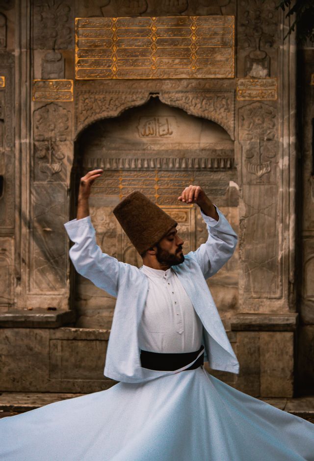 Un hombre con túnicas blancas tradicionales y un sombrero alto marrón realiza una danza giratoria frente a un muro de piedra ornamentado y tallado.