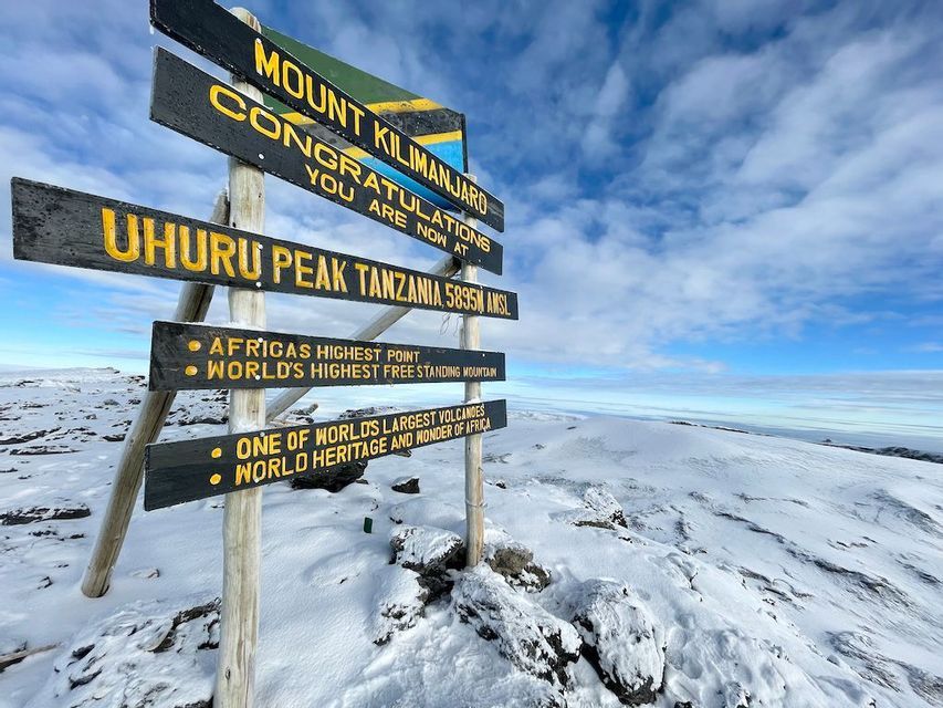L'iconico palo segnaletico a Uhuru Peak, sul Monte Kilimanjaro, eretto sulla cima innevata sotto un cielo azzurro e nuvoloso.