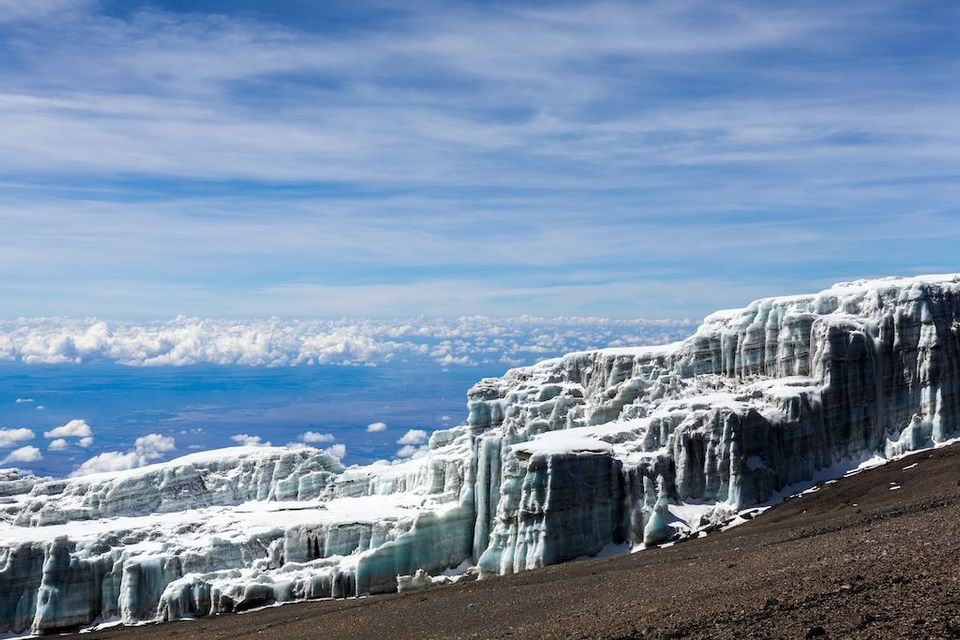 Ampia vista di un ghiacciaio su un fianco roccioso di montagna, con un mare di nuvole che si estende all'orizzonte sotto un cielo blu.