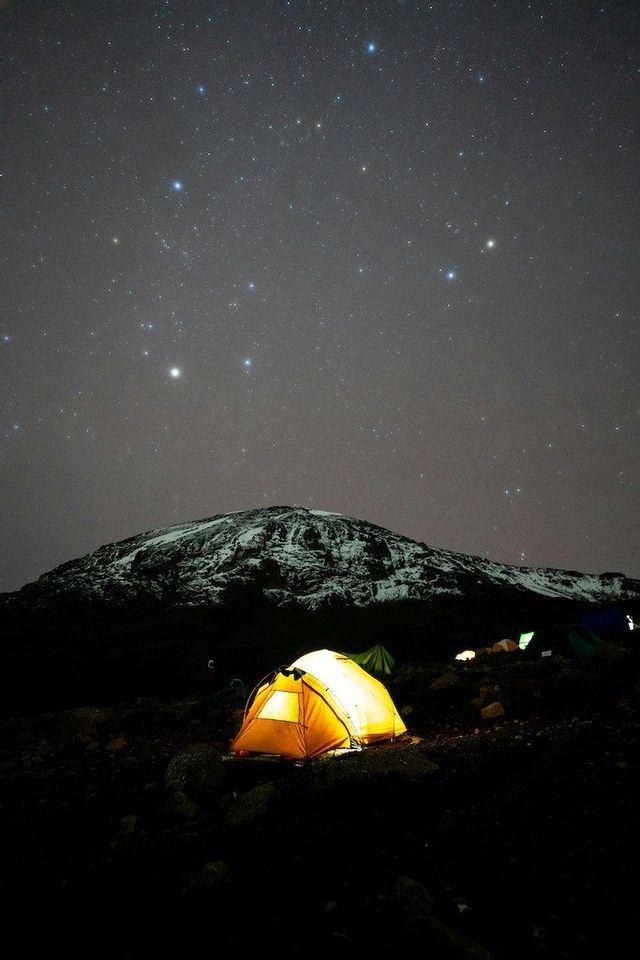 Una tenda illuminata si trova ai piedi di una montagna innevata sotto un cielo notturno pieno di stelle.