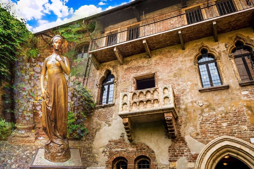 Una statua di bronzo di una donna si erge in un cortile acciottolato di fronte a un edificio rustico in mattoni con un balcone in pietra ornato.