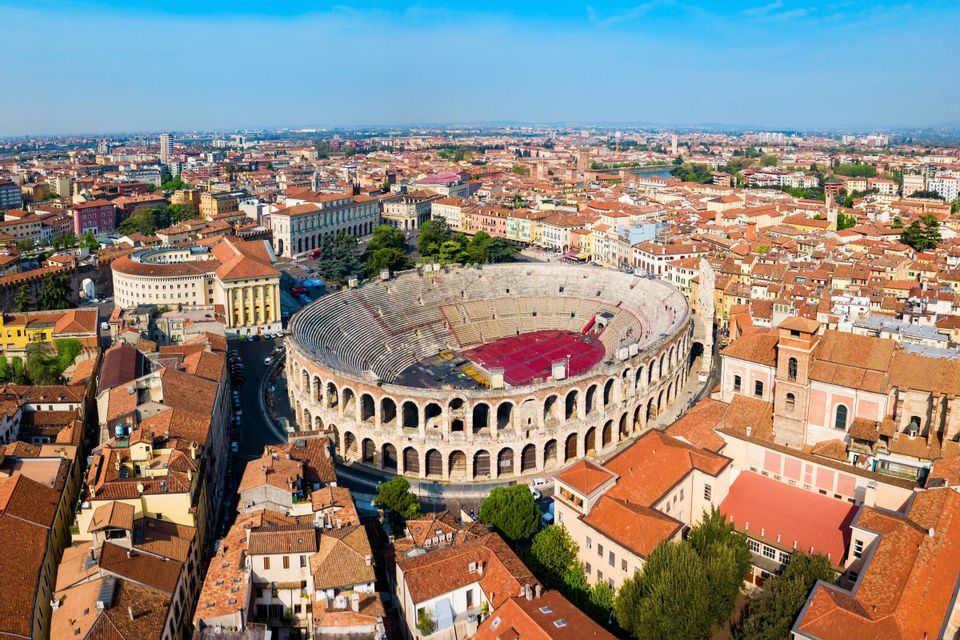 Vista aerea di un grande anfiteatro in pietra, circondato da una città con tetti rossi sotto un cielo azzurro e limpido.
