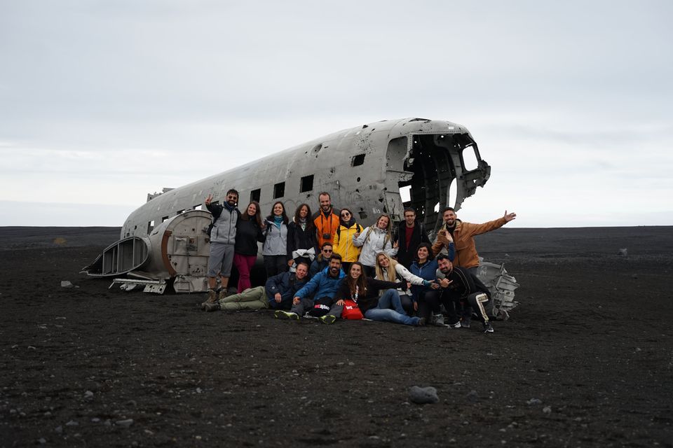 Un groupe WeRoad pose pour une photo devant un avion échoué sur une plage de sable noir sous un ciel nuageux.