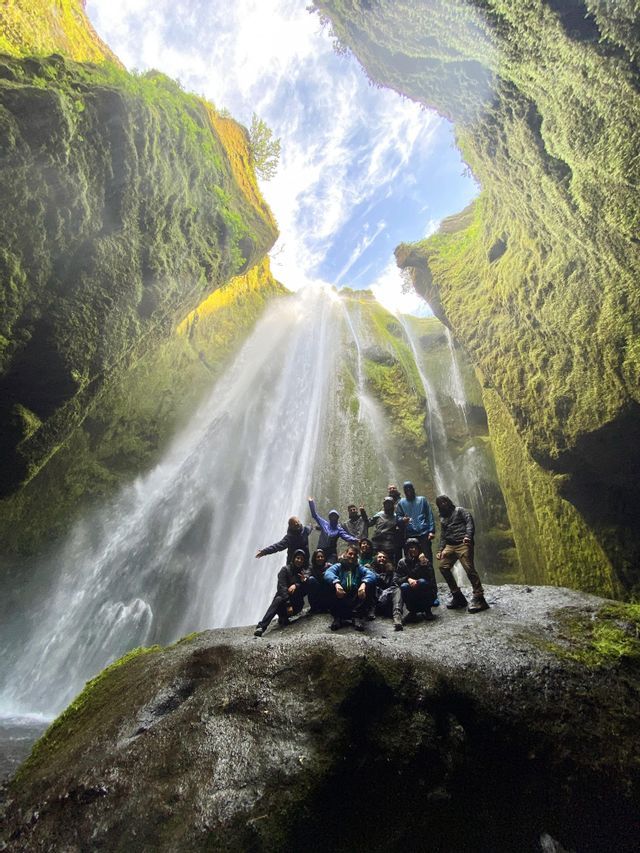 A WeRoad group trip poses on a large rock in front of a powerful waterfall inside a mossy green canyon.