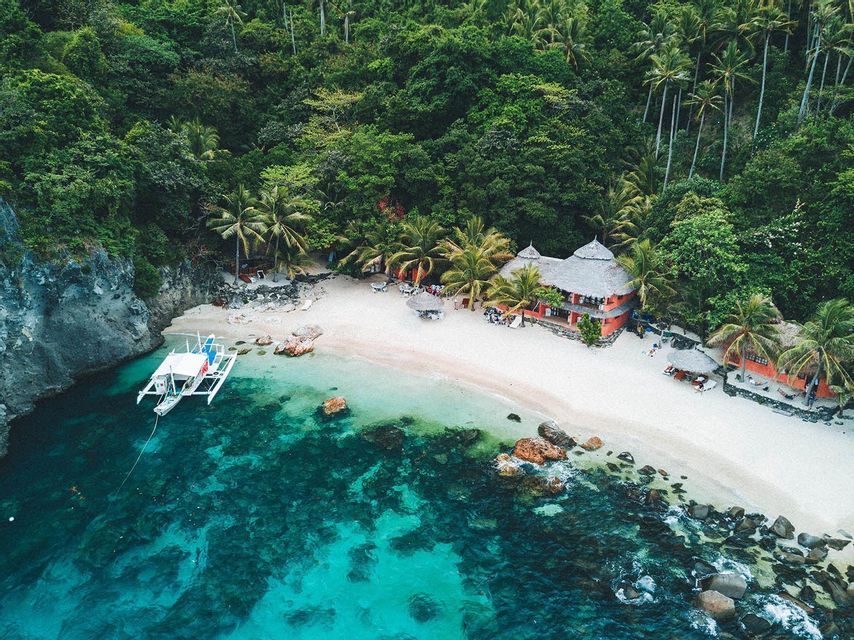 An aerial view of a secluded white sand beach with a thatched-roof building, bordered by a dense forest and clear turquoise water with a moored catamaran.