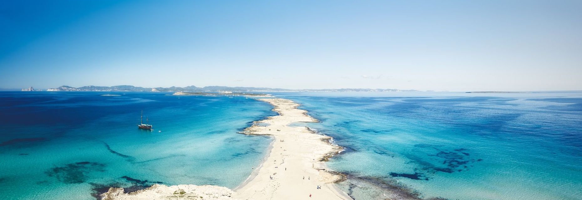 Vue aérienne d'une étroite langue de sable s'étendant dans une mer turquoise, avec un voilier ancré au large sous un ciel bleu clair.
