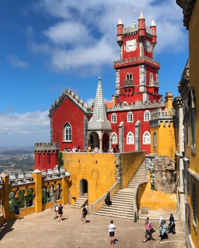 Menschen erkunden den Hof eines leuchtend rot-gelben Schlosses mit einem markanten Uhrturm unter einem teils bewölkten Himmel.