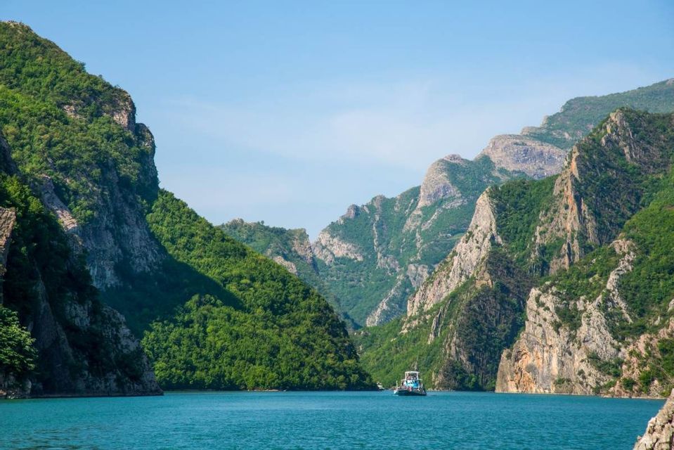 Un traghetto naviga su acque calme e turchesi attraverso un canyon di montagne ripide e verdi sotto un cielo sereno.