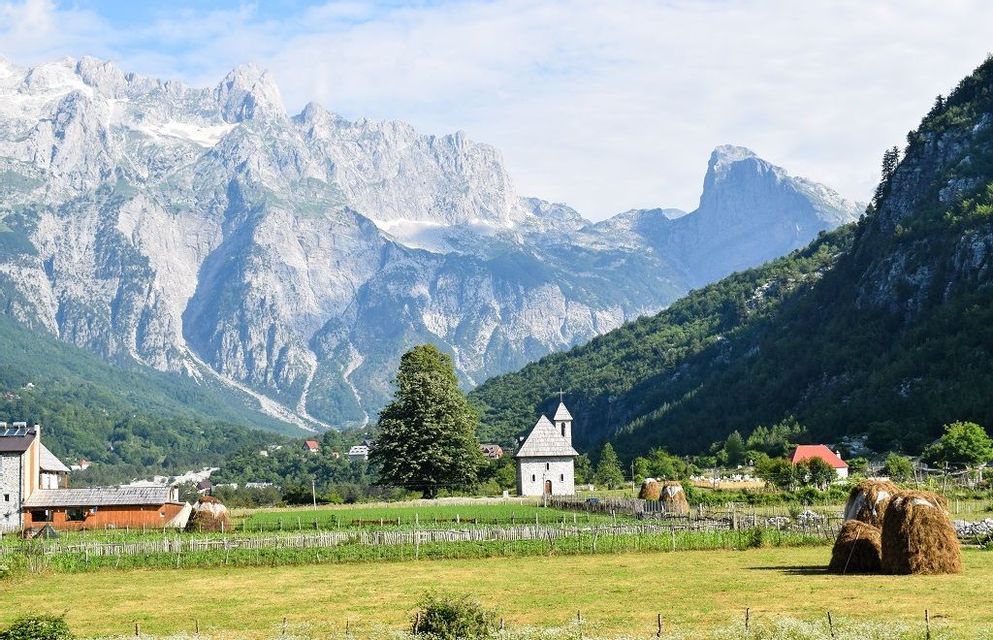 Una piccola chiesa in pietra e diversi covoni di fieno si trovano in un campo verde in una valle, con uno sfondo di grandi montagne aspre.