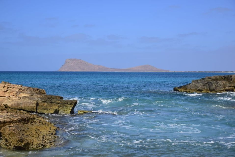Una vista desde una costa rocosa de un océano turquesa con una isla desierta en el horizonte bajo un cielo azul claro.
