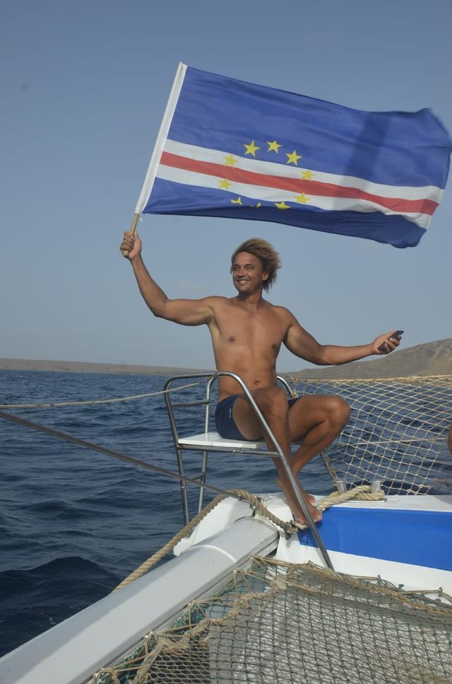 Un hombre rubio sonriente y sin camiseta se sienta en la proa de un barco, sosteniendo la bandera de Cabo Verde en alto sobre el agua.