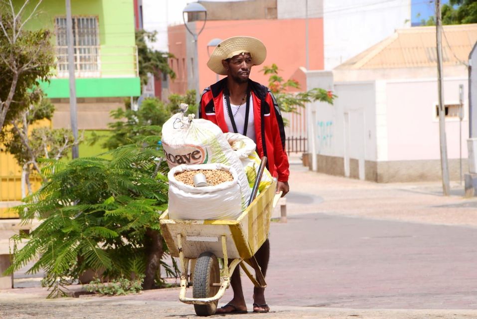 Un hombre con sombrero de paja empuja una carretilla llena de sacos de grano en una calle soleada con edificios coloridos al fondo.