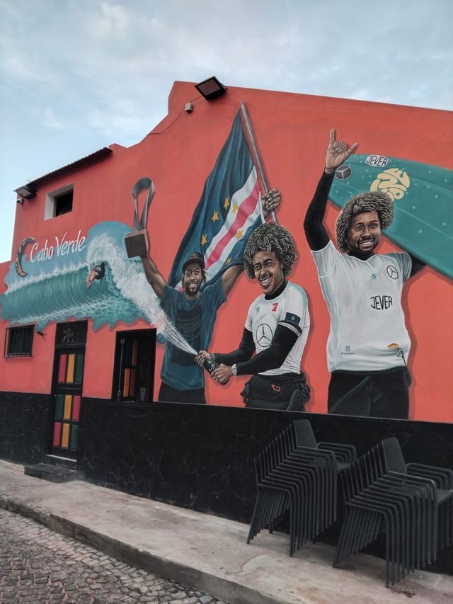 Un gran mural en la pared de un edificio naranja representa a atletas celebrando con un trofeo, una tabla de surf y la bandera de Cabo Verde.
