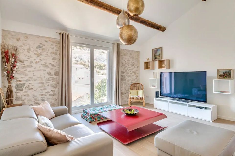 Un salon moderne avec des murs en pierres apparentes, des poutres en bois au plafond, un canapé clair et une table basse rouge.