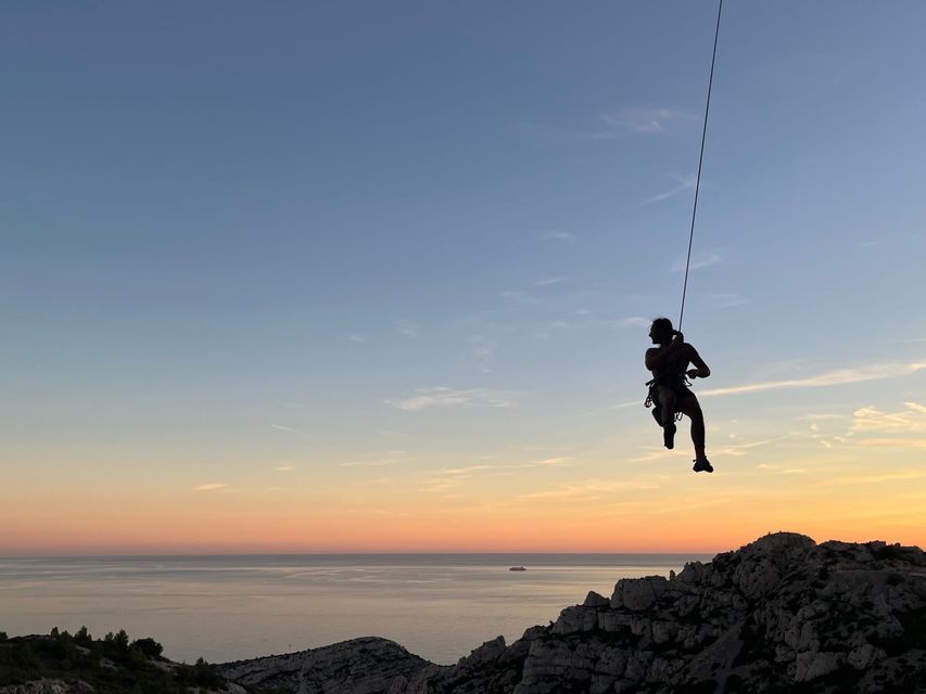 Une personne en silhouette dans un harnais, suspendue à une corde au-dessus de falaises rocheuses et de la mer au coucher du soleil.