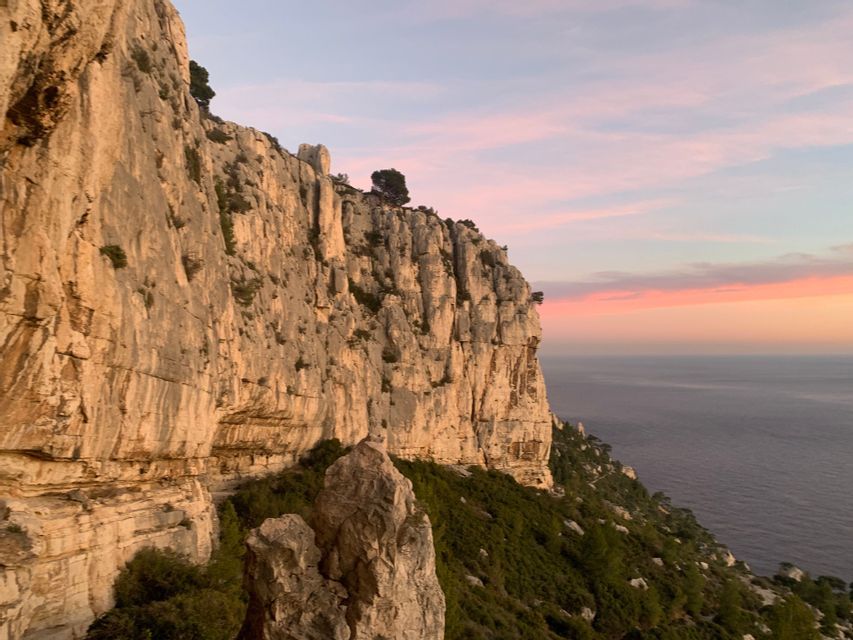 Une falaise escarpée et rocheuse avec des arbustes verts surplombe la mer calme lors d'un coucher de soleil coloré.