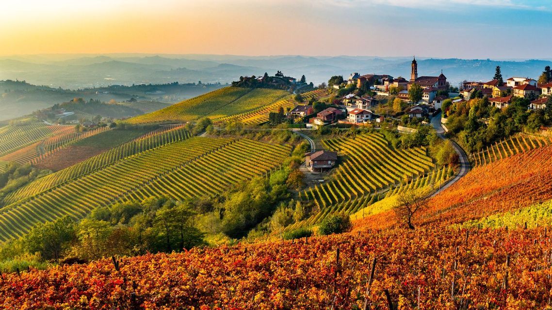 Un borgo sorge su una collina che domina dolci colline coperte di vigneti dai vivaci colori autunnali all'alba.