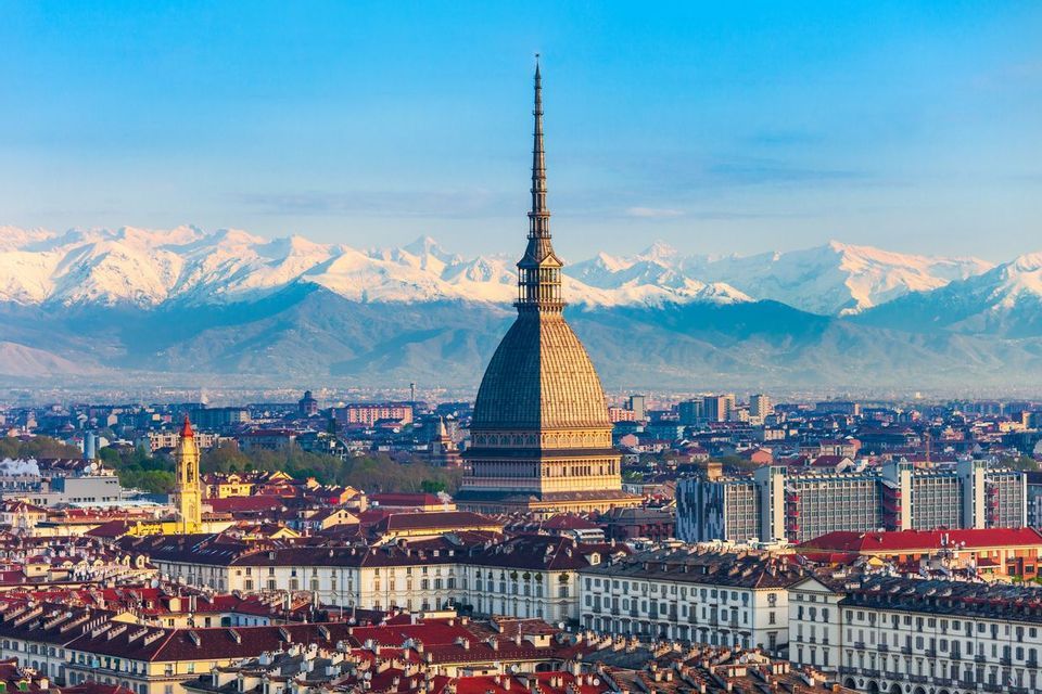 La cupola e la guglia della Mole Antonelliana si ergono sopra un paesaggio urbano con uno sfondo di montagne innevate sotto un cielo azzurro.