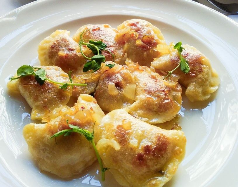 Un primo piano di pierogi saltati in padella su un piatto bianco, guarniti con cipolle saltate e micro-germogli verdi freschi.