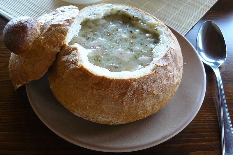 Una zuppa cremosa servita all'interno di una ciotola di pane svuotata con il suo coperchio, posizionata su un piatto accanto a un cucchiaio.