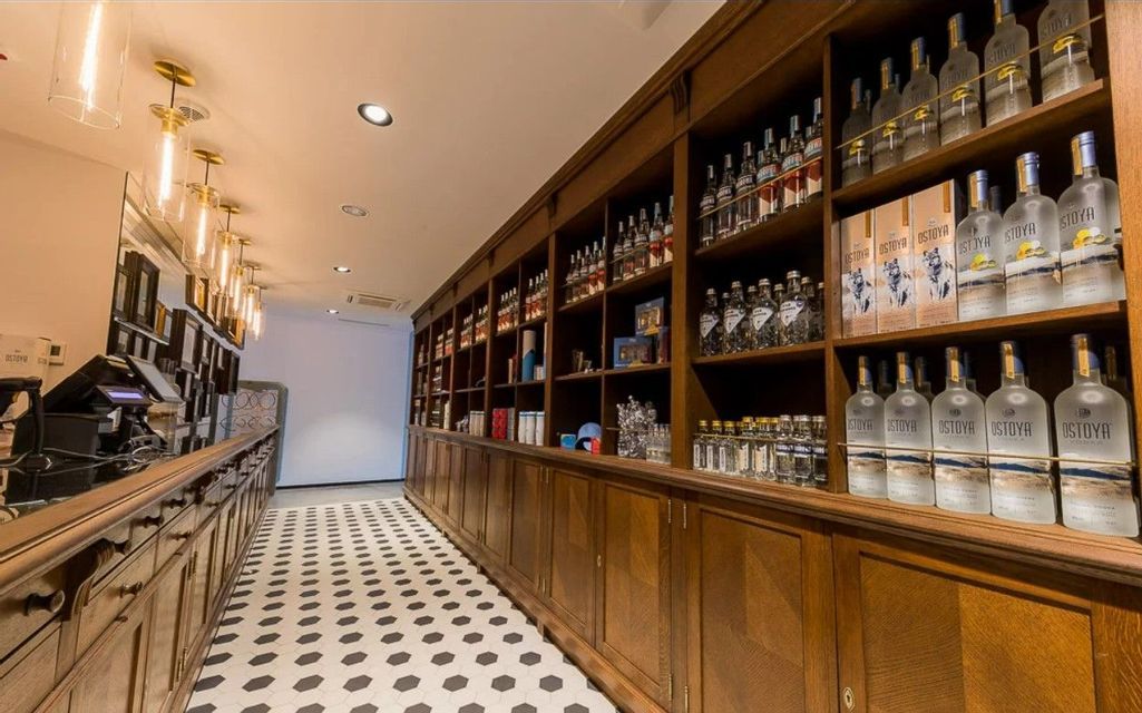 Interno di un negozio di liquori con scaffali in legno ricolmi di bottiglie di vodka Ostoya e un pavimento a piastrelle geometriche.