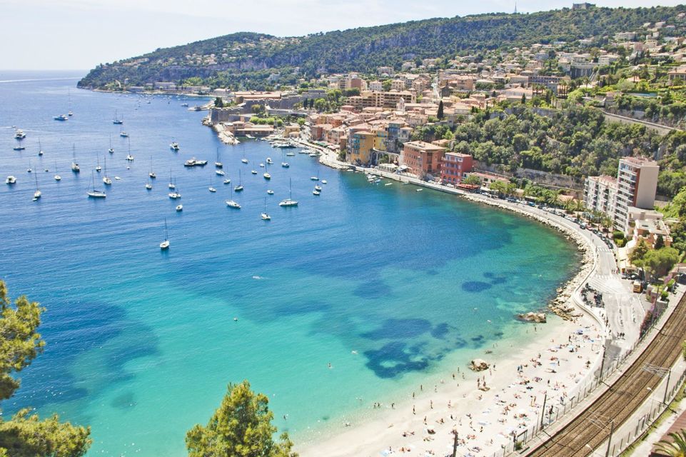 An aerial view of a coastal town with a sandy beach full of people and a bay with many sailboats anchored in turquoise water.
