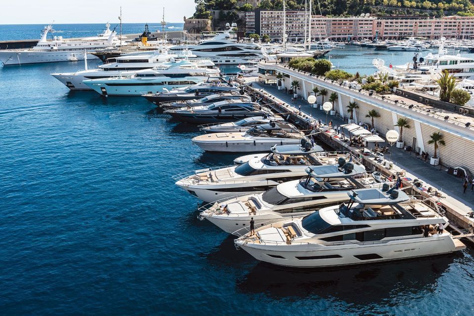Numerous luxury yachts and boats are docked in rows at a pier in a large, sunny harbor.