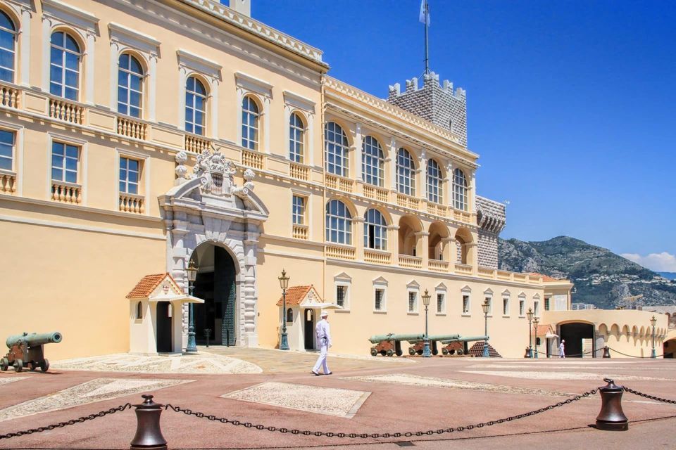 A guard in a white uniform walks across the courtyard of a grand palace lined with cannons under a clear blue sky.