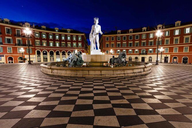 A fountain with a tall white statue stands in a square with checkerboard paving, surrounded by illuminated buildings at night.