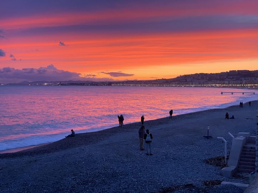 Silhouettes of people on a pebble beach watching a vibrant orange and purple sunset over the sea, with a coastline in the background.