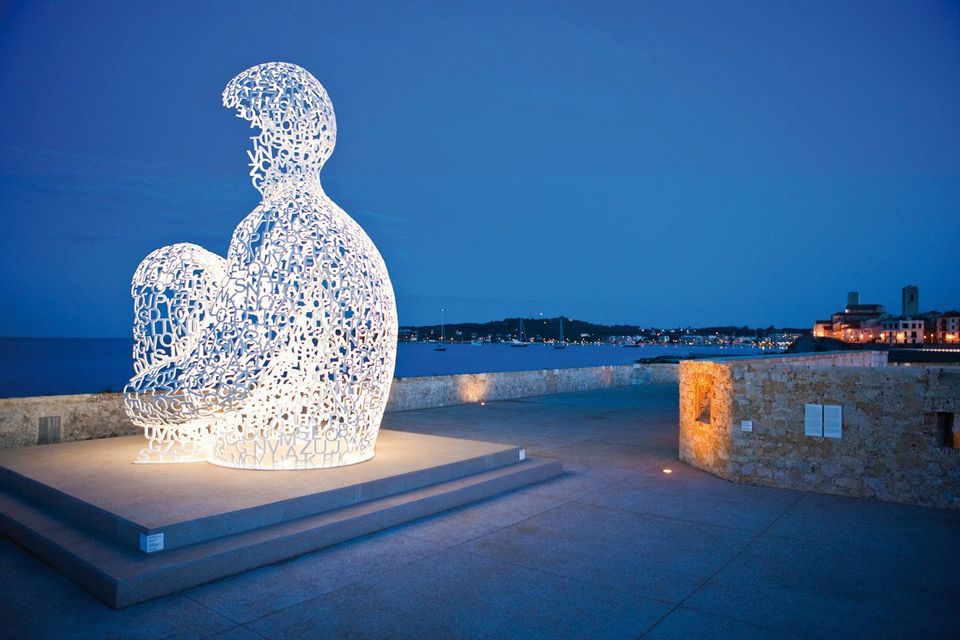 An illuminated white sculpture made of letters sits on a stone promenade overlooking the sea at dusk.