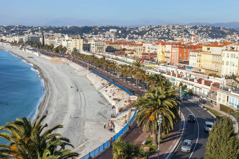 An aerial view of a coastal city with a long pebble beach, a palm-tree-lined promenade, and colorful buildings under a clear sky.