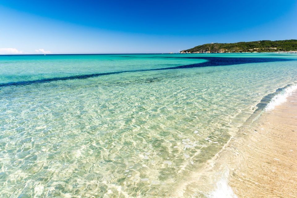 Clear turquoise water laps against a sandy beach with a green headland in the background under a blue sky.
