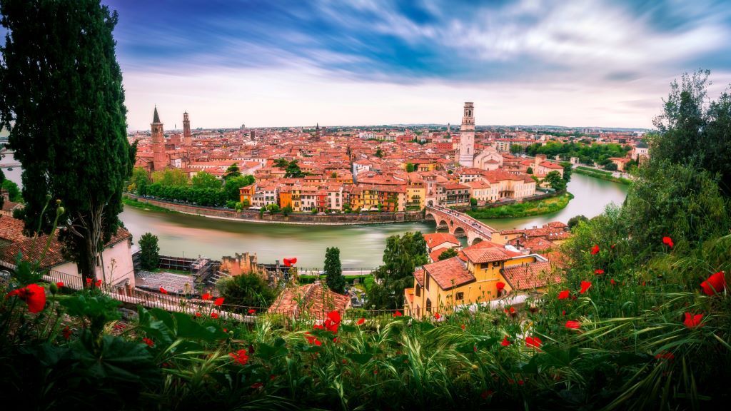 Un ampio paesaggio urbano di edifici dai tetti rossi lungo un fiume sinuoso con un ponte ad arco in pietra, visto da una collina con fiori rossi.