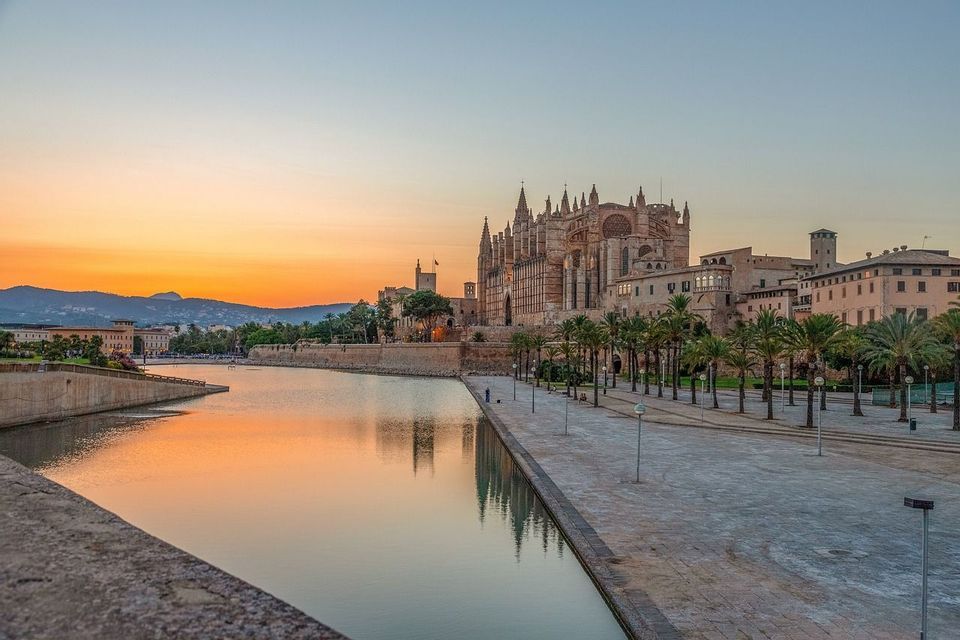 Eine große Kathedrale spiegelt sich bei Sonnenuntergang in einem Gewässer, neben einer breiten, von Palmen gesäumten Promenade.