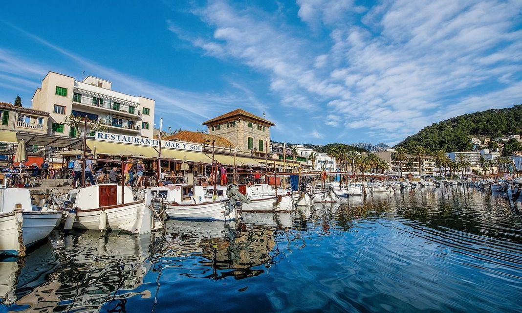 Kleine weiße Boote liegen in einem Hafen vor Uferrestaurants und Gebäuden unter blauem Himmel.
