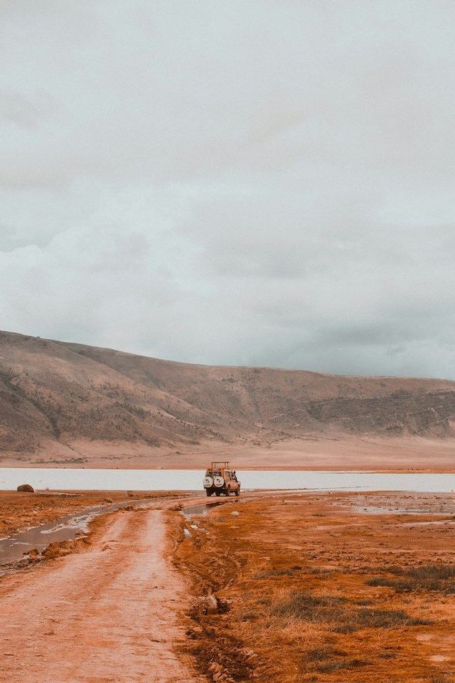 Una jeep safari percorre una strada sterrata accanto a uno specchio d'acqua, con una grande collina sullo sfondo sotto un cielo nuvoloso.