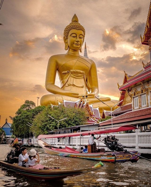 Des bateaux à longue queue naviguent sur un canal, avec une statue géante de Bouddha doré et un temple en arrière-plan, au coucher du soleil.