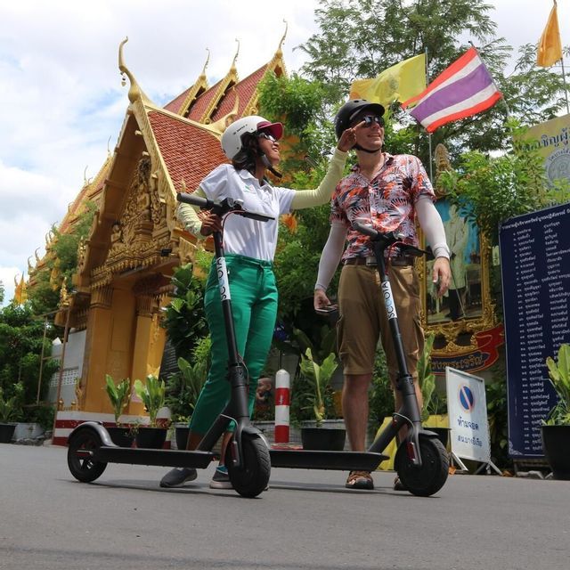 Une femme ajuste le casque d'un homme alors qu'ils se tiennent avec des trottinettes électriques devant un temple doré orné d'un toit de tuiles rouges.