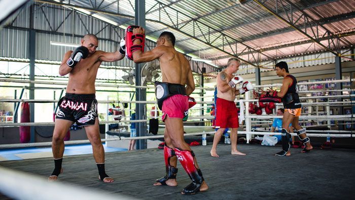 Des hommes d'un voyage de groupe WeRoad lors d'une leçon de Muay Thai, s'entraînant aux coups de poing avec des entraîneurs sur un ring de boxe.