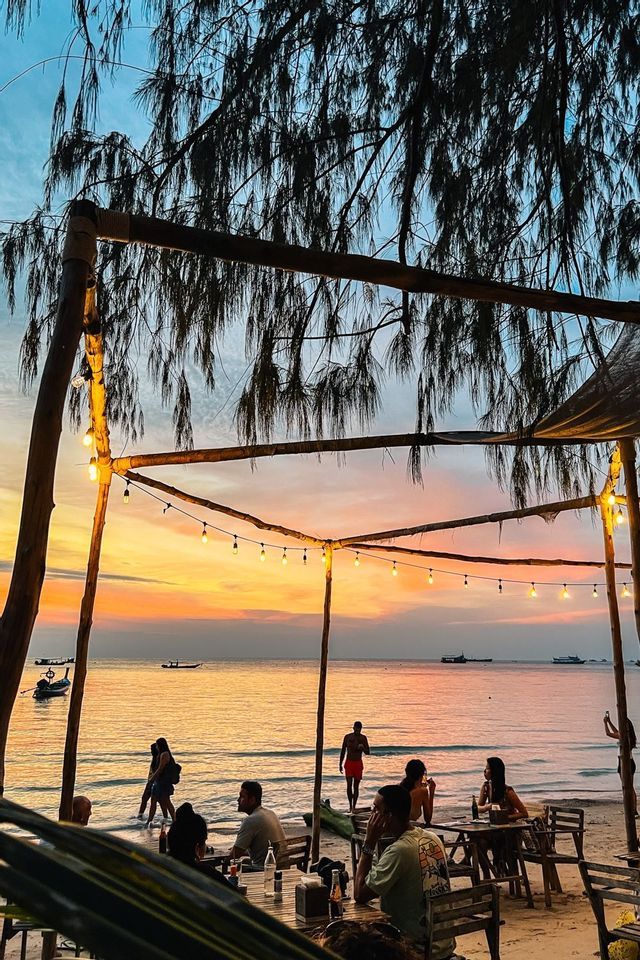 Un groupe WeRoad se détend dans un bar de plage sous des guirlandes lumineuses, regardant le coucher de soleil sur la mer.
