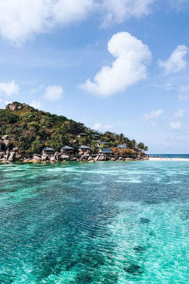 Une île verte et rocheuse avec des bungalows le long du rivage, entourée d'une eau turquoise claire sous un ciel partiellement nuageux.