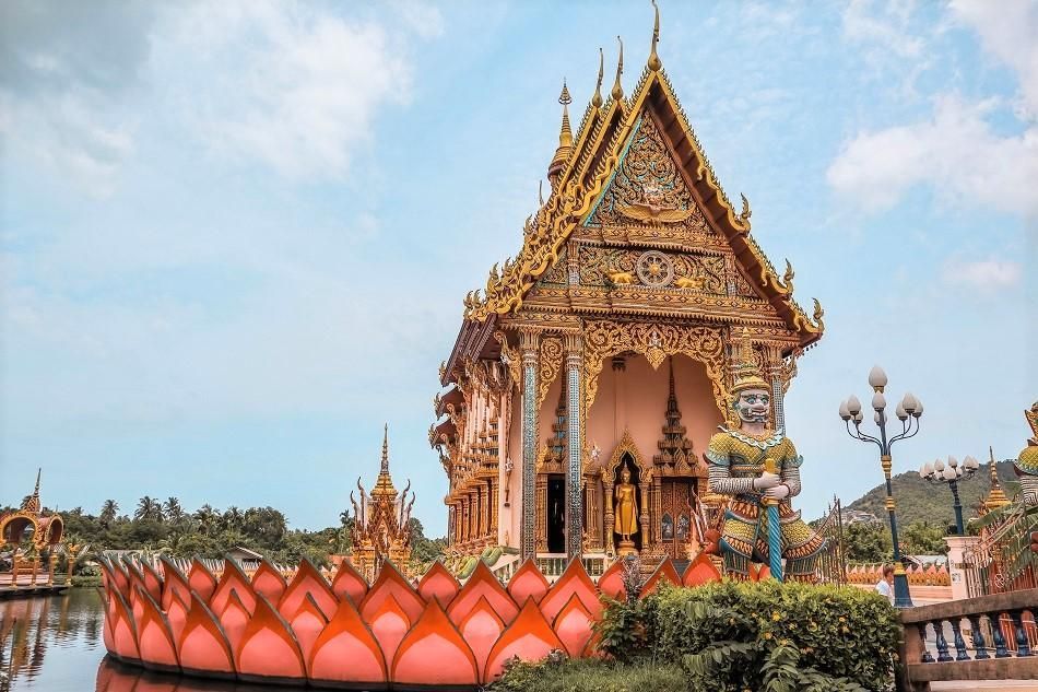 Un temple doré orné avec une statue gardienne à côté d'un étang et une grande fleur de lotus rose.