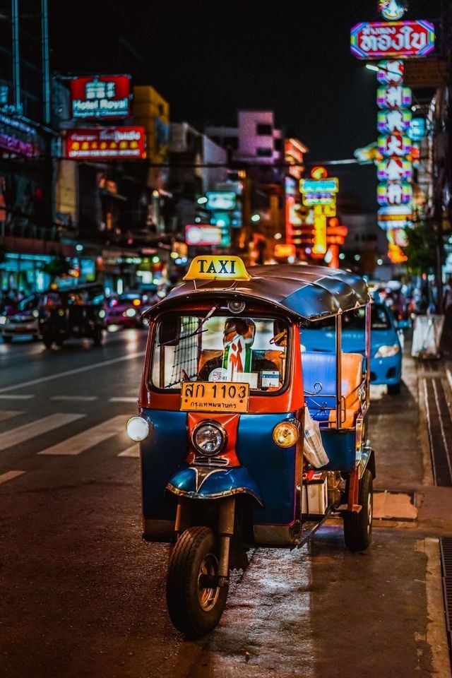 Un taxi tuk-tuk rouge et bleu est garé dans une rue de ville mouillée la nuit, avec des néons colorés illuminant l'arrière-plan.
