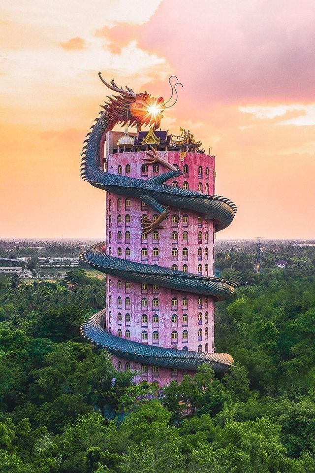 Une grande sculpture de dragon vert s'enroule autour d'un grand bâtiment cylindrique rose dans une forêt luxuriante au coucher du soleil coloré.