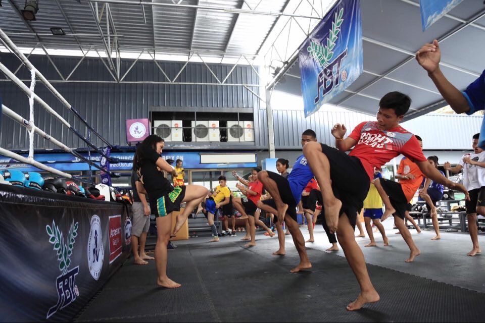 Un voyage de groupe WeRoad avec des participants s'entraînant aux high kicks dans une salle de Muay Thai, à côté d'un ring de boxe.