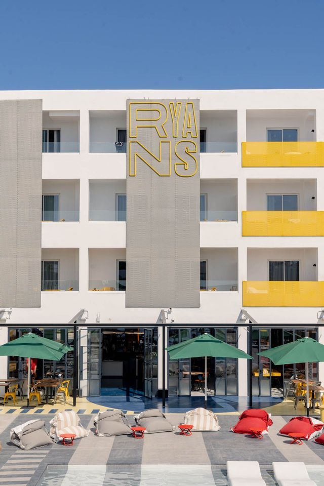 The modern white facade of the Ryans hotel, with a poolside patio featuring bean bag chairs and green umbrellas under a clear sky.