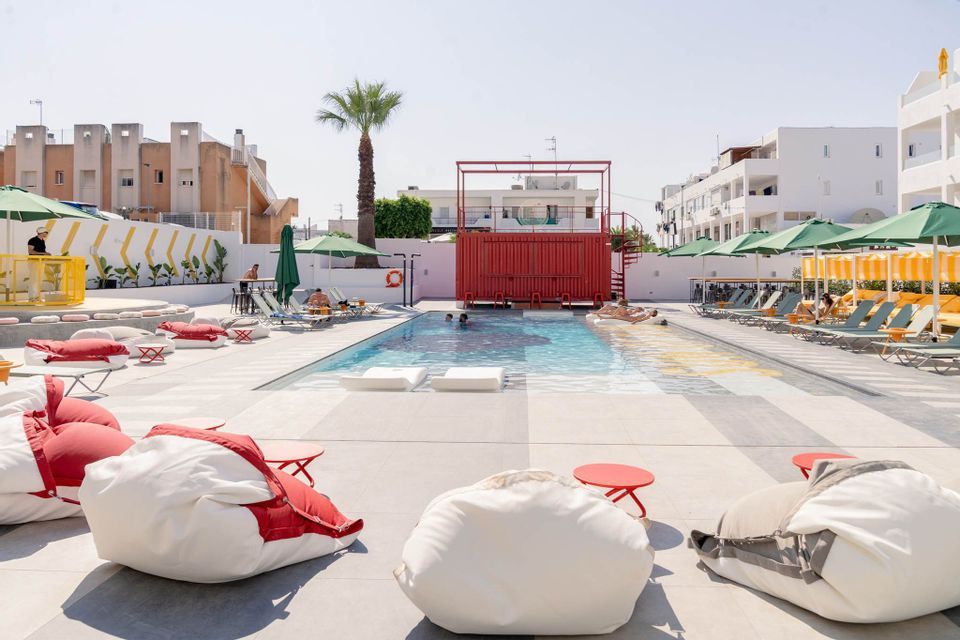 Una vista soleggiata di una piscina d'hotel con persone che nuotano, circondata da lettini, pouf e un container rosso.