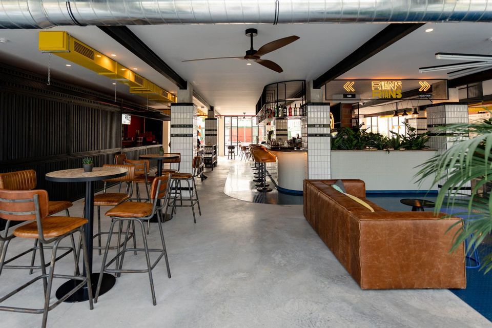 The empty interior of a modern industrial-style bar with high-top tables, brown leather stools, and a long leather sofa.